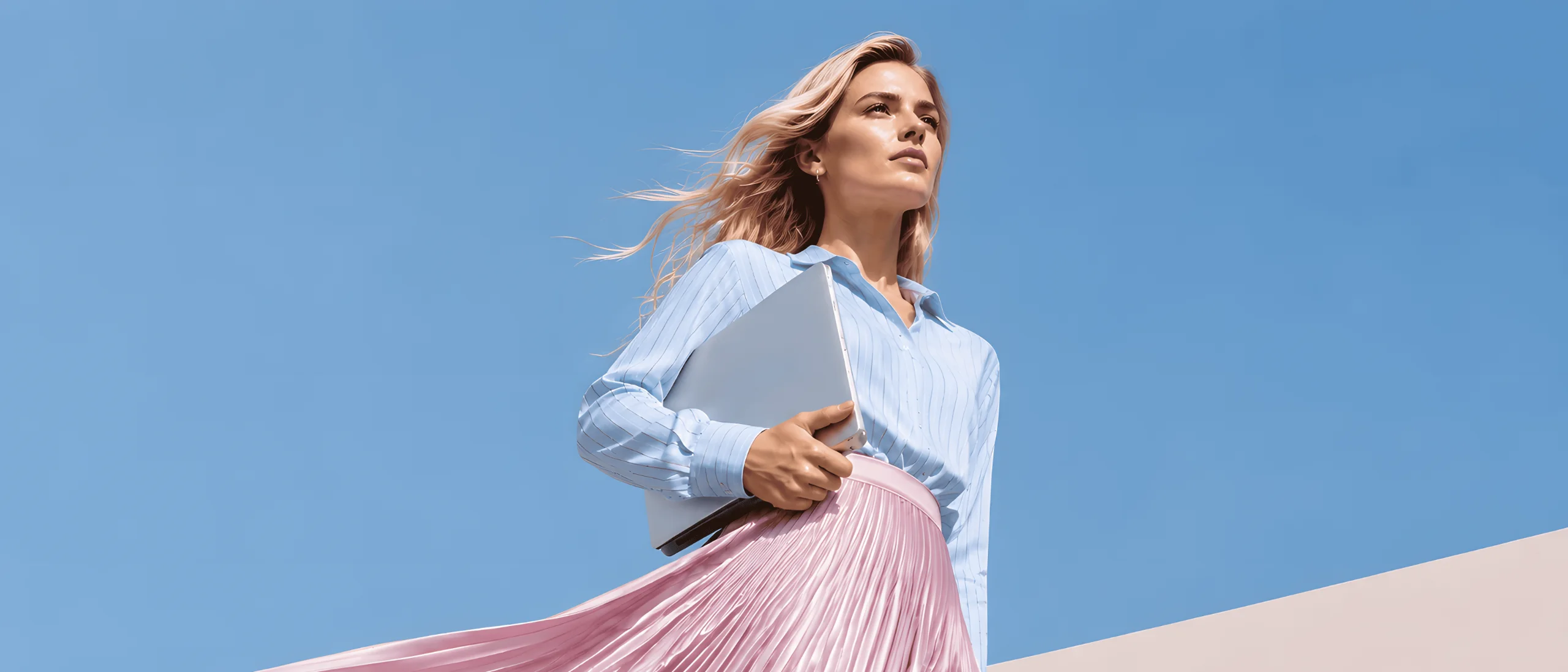 A woman in a flowing pleated skirt holding a laptop against a clear blue sky